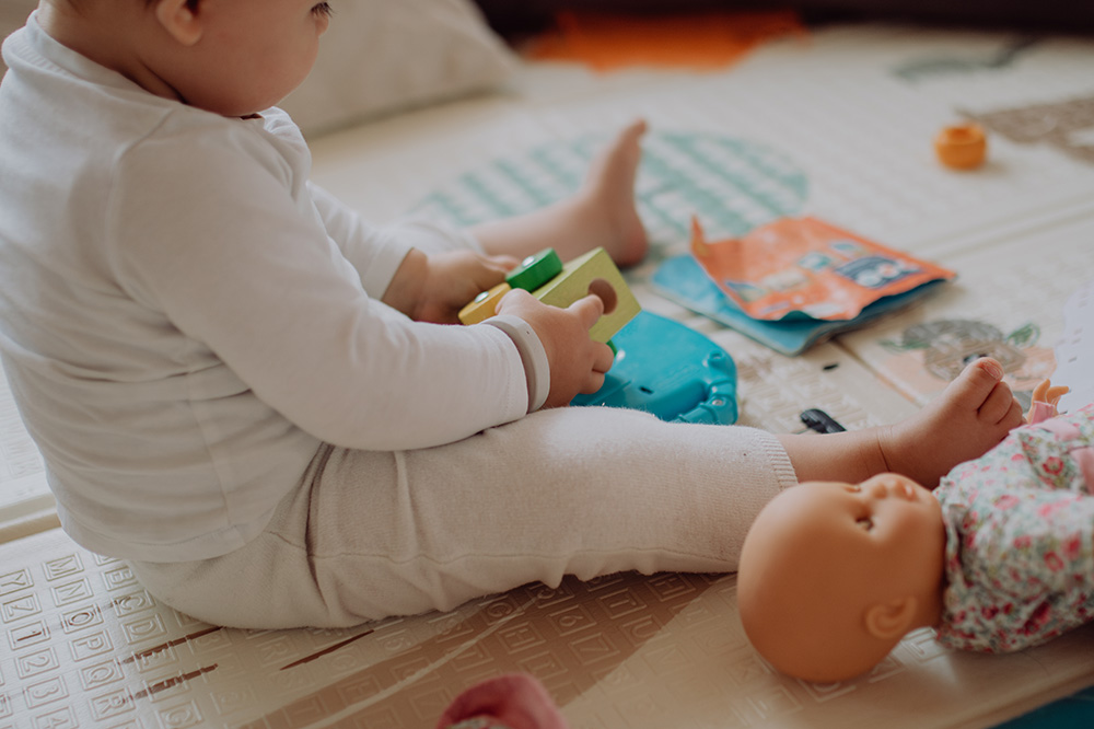 Baby sitzt auf einer gepolsterten Spielmatte und hält buntes Holzspielzeug in den Händen. Vor dem Baby liegt eine Babypuppe in einem geblümten Outfit. Daneben befinden sich ein offenes Bilderbuch und weiteres Spielzeug. Im Hintergrund ist ein orangefarbenes Spielobjekt zu erkennen.