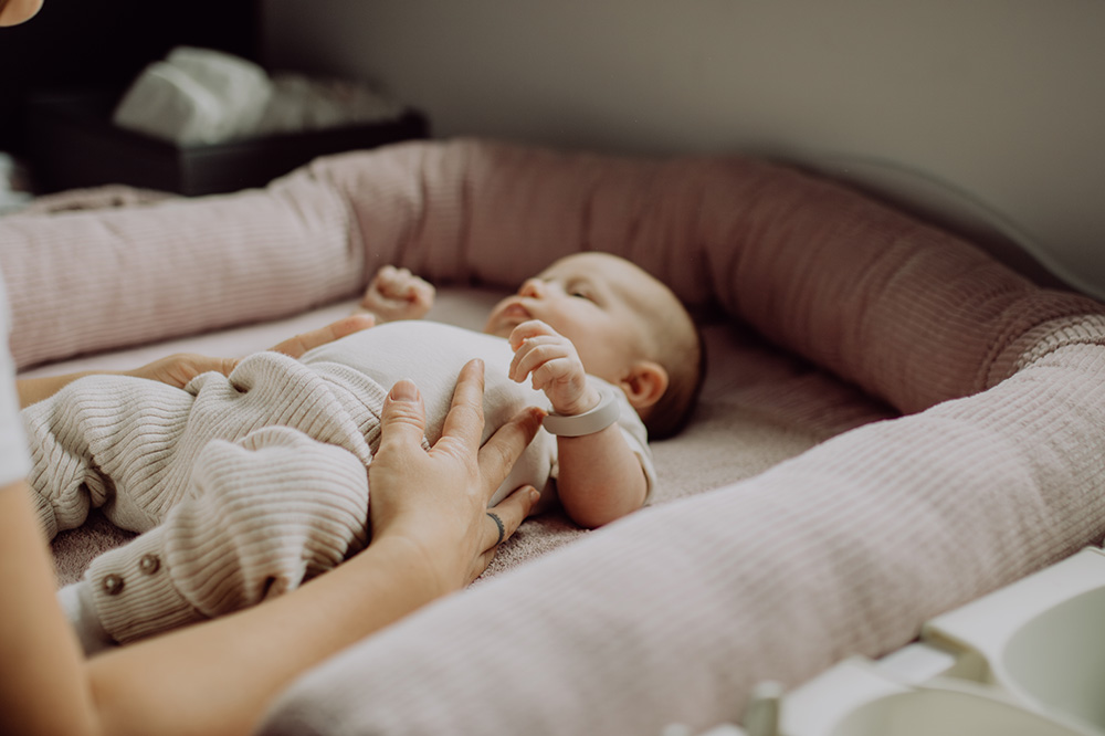 Baby liegt auf einer Wickelkommode in einem weich gepolsterten Nest mit rosa Stoff. Eine Hand eines Erwachsenen hält sanft das Baby am Bauch, während das Baby entspannt auf dem Rücken liegt. Im Hintergrund ist eine schwarze Box mit Pflegeartikeln zu erkennen.