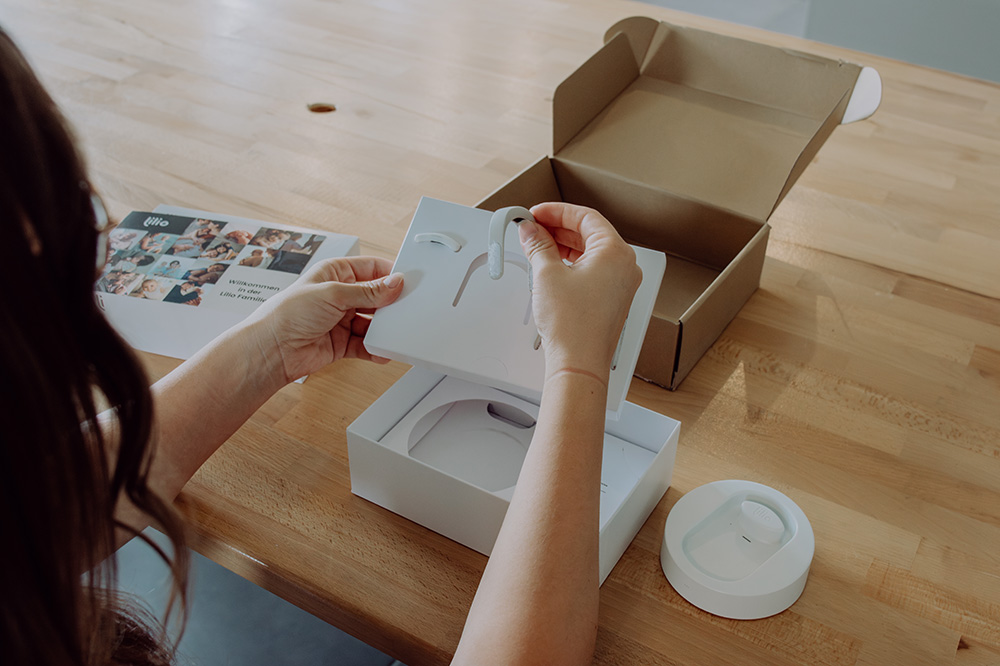 Frau packt lilio armband aus. Eine Person packt ein technisches Gerät aus einer weißen Box aus. Sie hält ein Teil des Produkts in der Hand, während die geöffnete Verpackung auf einem Holztisch liegt. Daneben befindet sich eine runde, weiße Ladestation. Im Hintergrund liegt eine Broschüre mit Bildern und Text. Eine braune Versandverpackung ist ebenfalls geöffnet auf dem Tisch zu sehen.